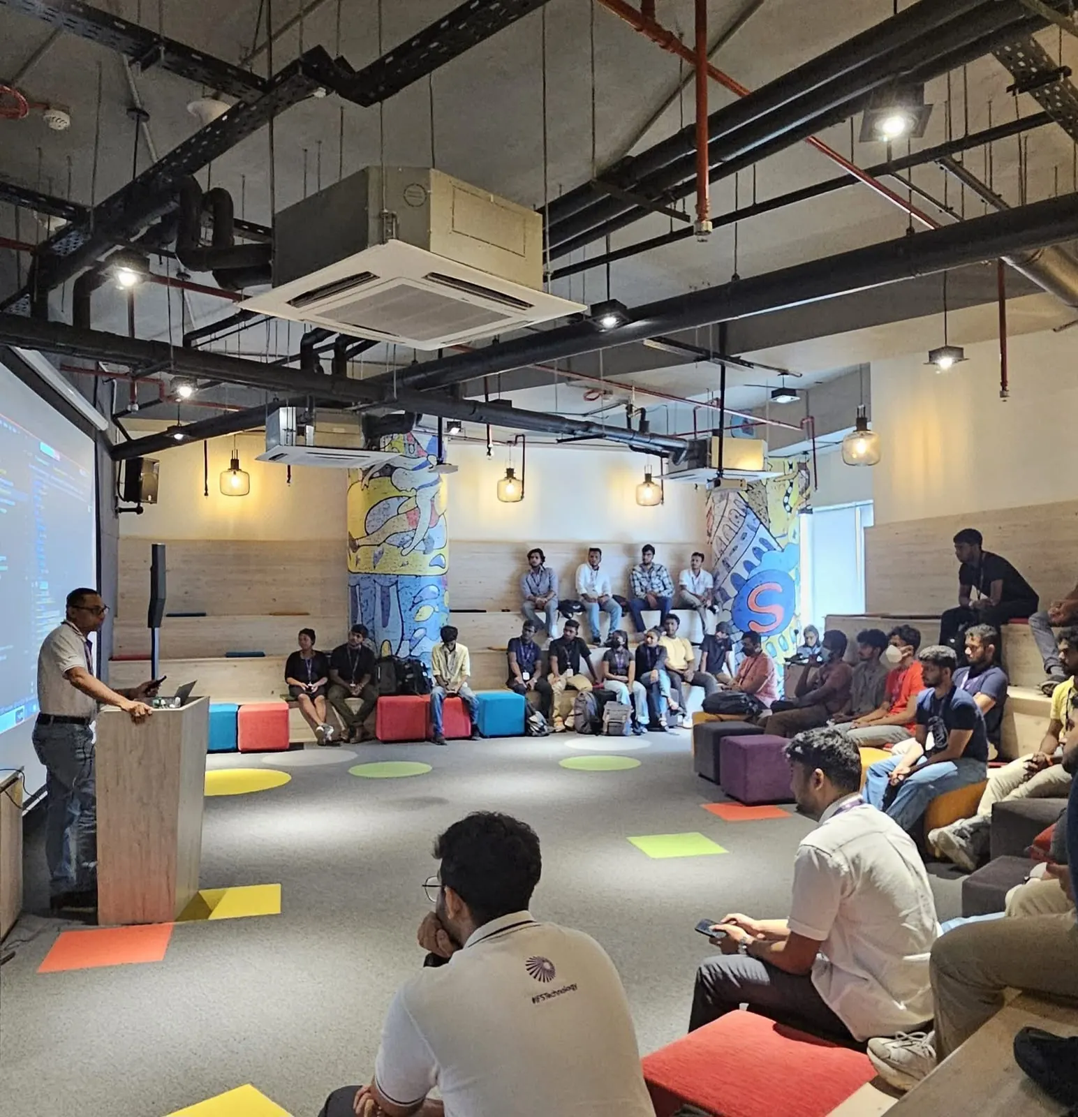 Speaker presenting at a podium to a seated audience in a colorful, modern venue